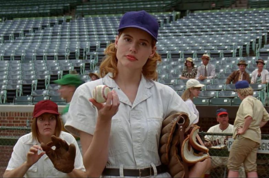 2 women in baseball uniforms, one in center holding a ball and mitt and one on the left with a mitt ready to catch